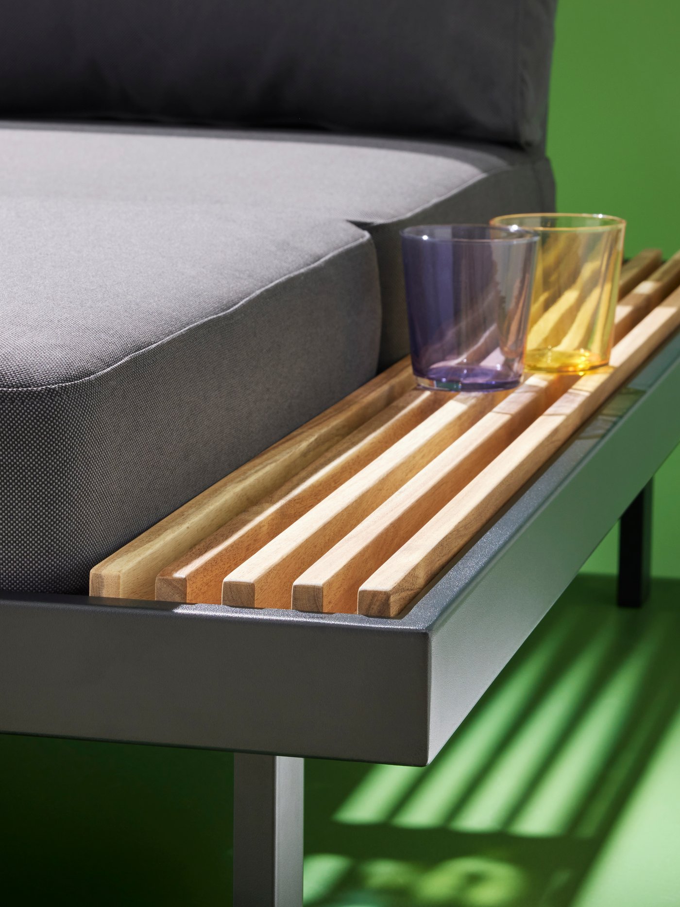 A yellow and a purple glass are resting on the integrated side table of a REVSKÄR 3-seat conversation set in a green space.