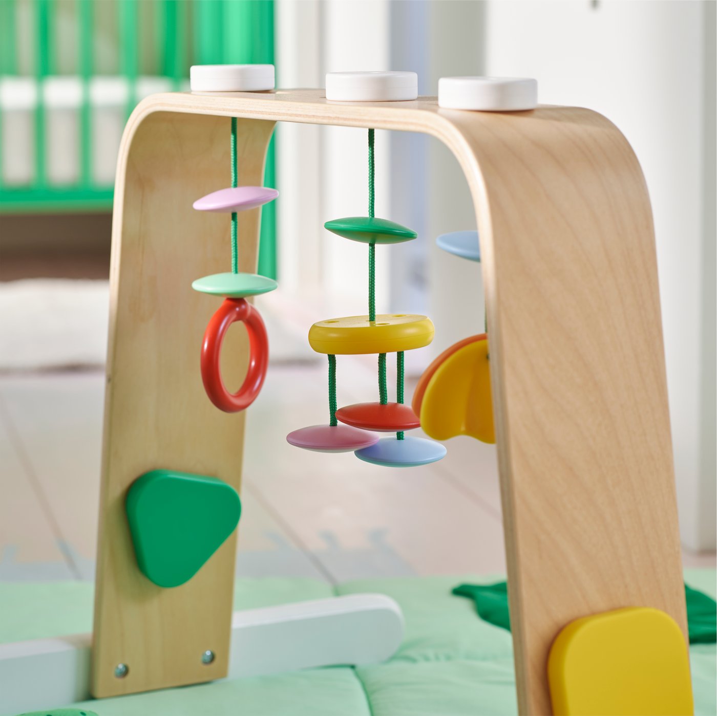 Un portique pour bébé LEKA avec des anneaux et des mobiles de couleurs vives sur un tapis de jeu vert dans une chambre d’enfant.