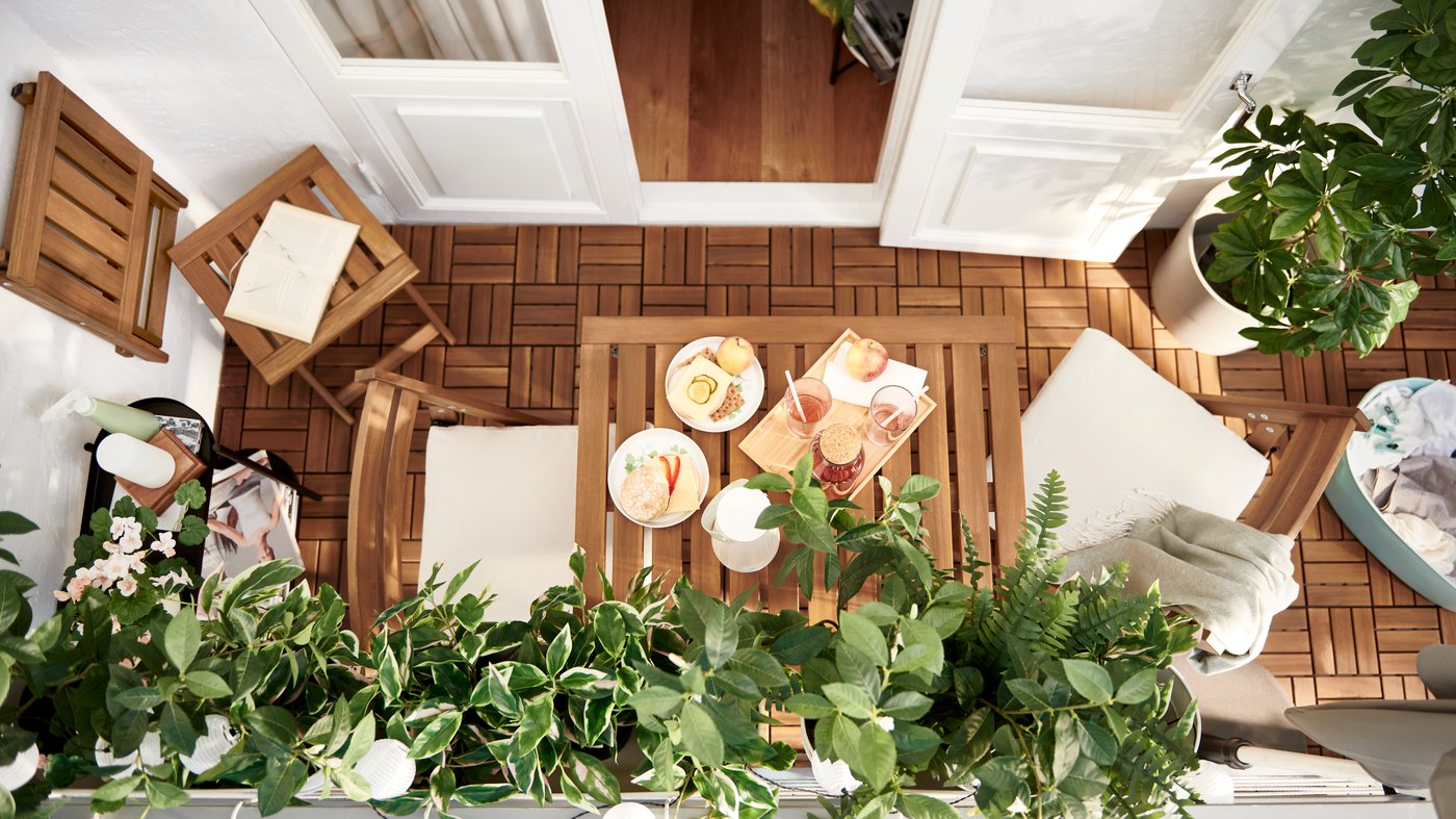 A balcony characterised by slatted acacia – NÄMMARÖ furniture and RUNNEN floor decking – and leafy FEJKA artificial plants.