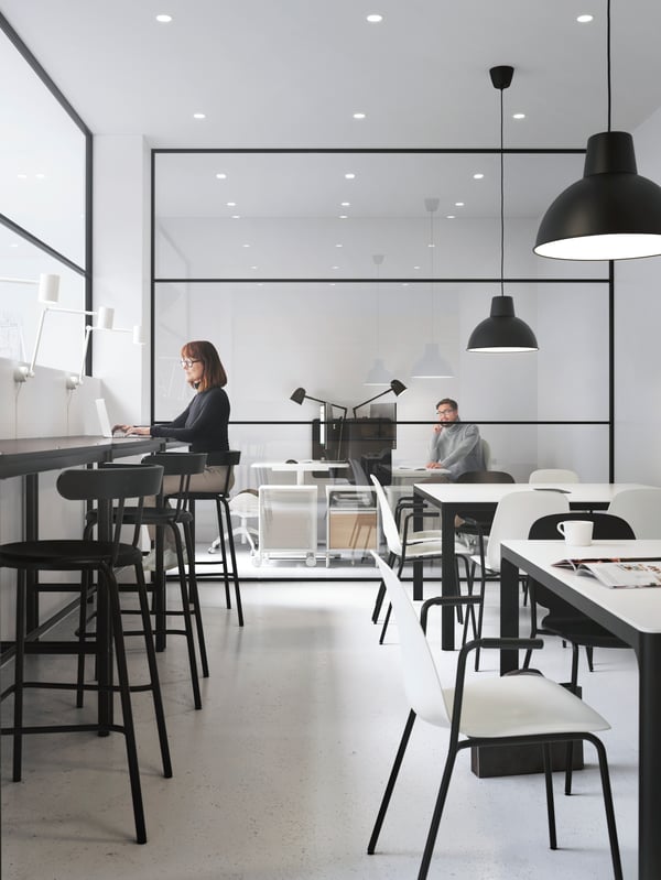 Two work areas, one bar-style with high stools, the other a row of tables. At the back, a man in a glass-fronted office.