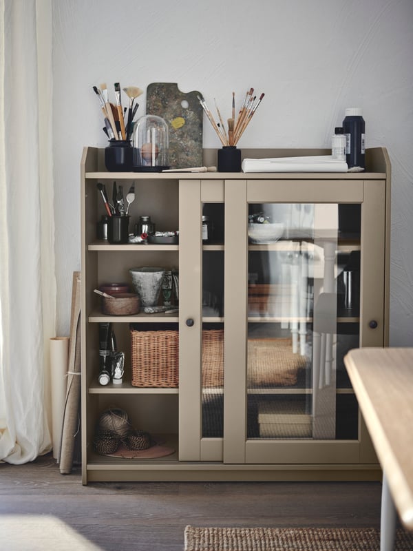 A wood-floor room with a beige HAUGA glass-door cabinet with painting accessories on top and miscellaneous storage inside.