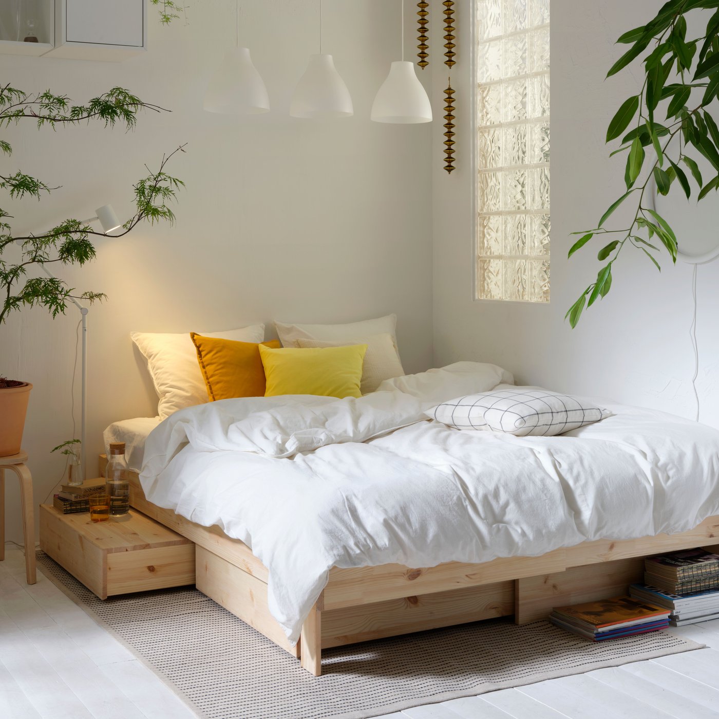 Es ist ein Schlafzimmer mit großem Fenster, einem GLAMBERGET Bettgestell aus unbehandeltem Holz mit Aufbewahrung, beigefarbener Bettwäsche und weißen PLATSA Schränken zu sehen.