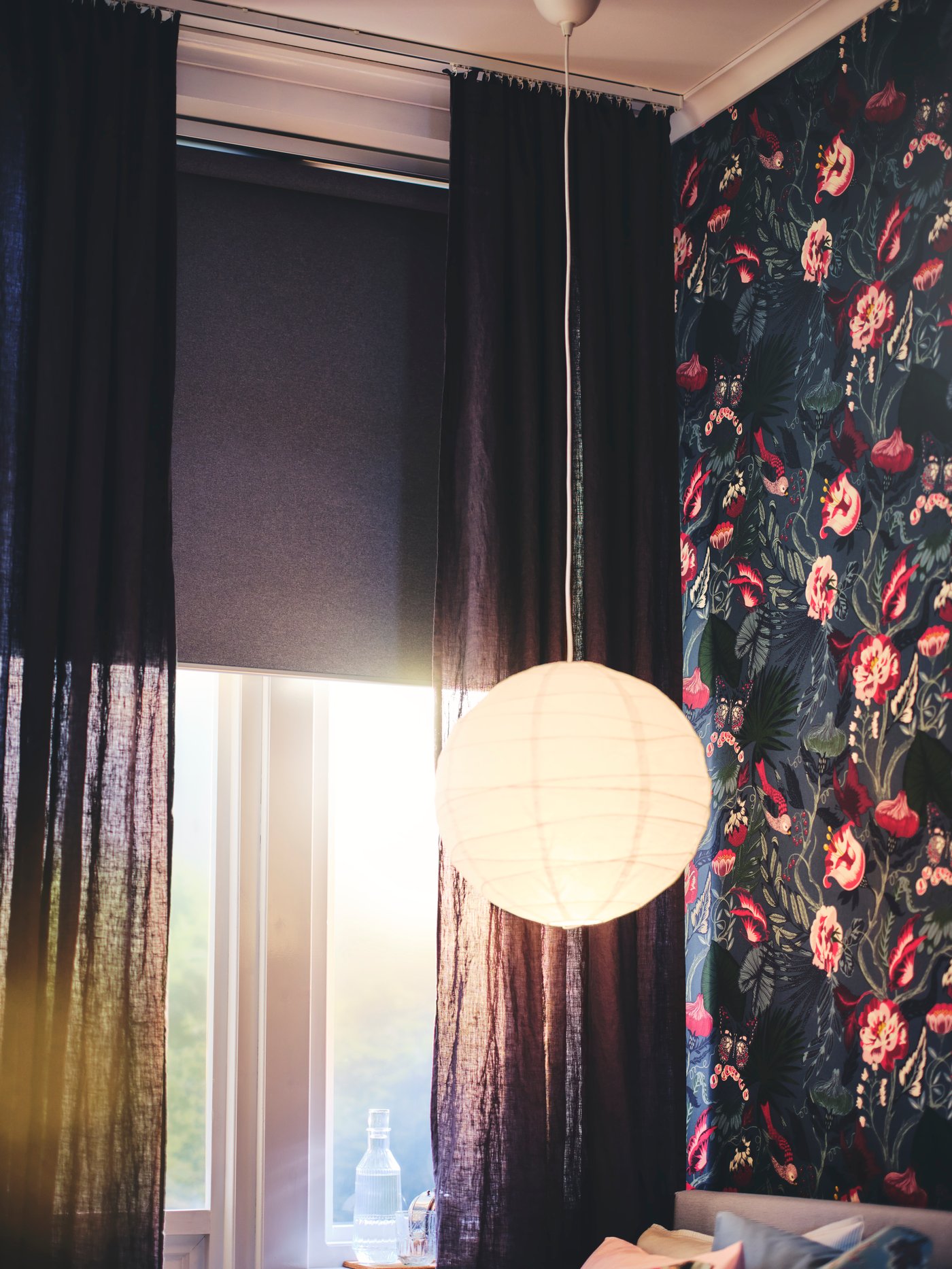 A lit pendant lamp in front of a window with a half-drawn SKOGSKLÖVER roller blind and a pair of DYTAG curtains.