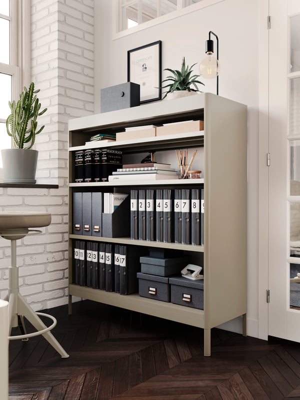 A white-brick-wall office interior with a full, neatly organised IDÅSEN shelving unit and a TROLLBERGET sit-stand support.