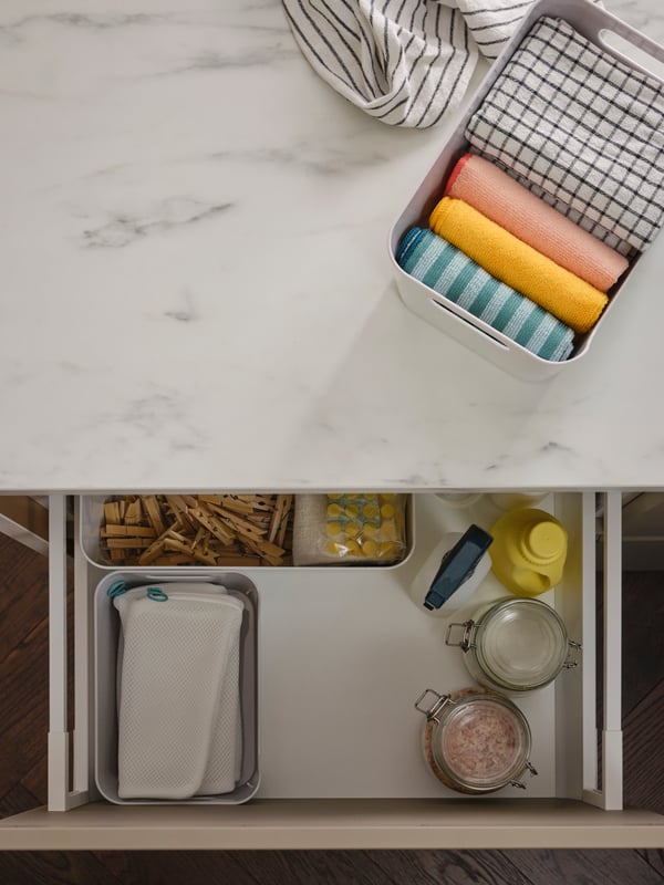 White VARIERA boxes holding laundry accessories inside a kitchen drawer, plus another box on a white marble effect worktop.