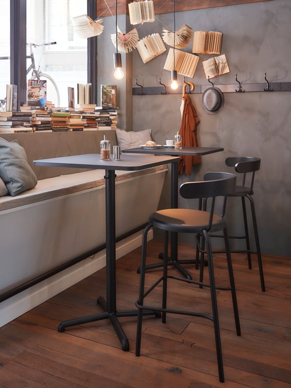 Two STENSELE bar tables in black with two YNGVAR bar chairs in a cosy corner of the living room with decorative bulbs and other items hung on the wall beside the bar tables.
