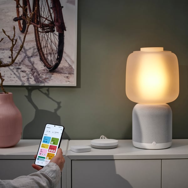 Bela SYMFONISK osnova lampe sa zvučnikom i staklenim abažurom stoji na ormariću. Ruka usmerava mobilni telefon prema njega.
