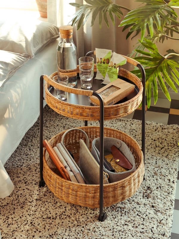 Ein praktischer, zweistöckiger Rattan-Trolley mit zwei Körben, ideal für die Aufbewahrung von Alltagsgegenständen. Auf der oberen Ablage befinden sich eine Wasserflasche, Gläser und ein Buch, während die untere Ebene Platz für Taschen und kleine Accessoires bietet.
