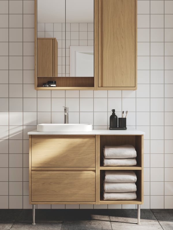 A white-tiled bathroom has an ÄNGSJÖN wash-stand in oak effect with legs, a semi-recessed wash-basin and a mirror cabinet.