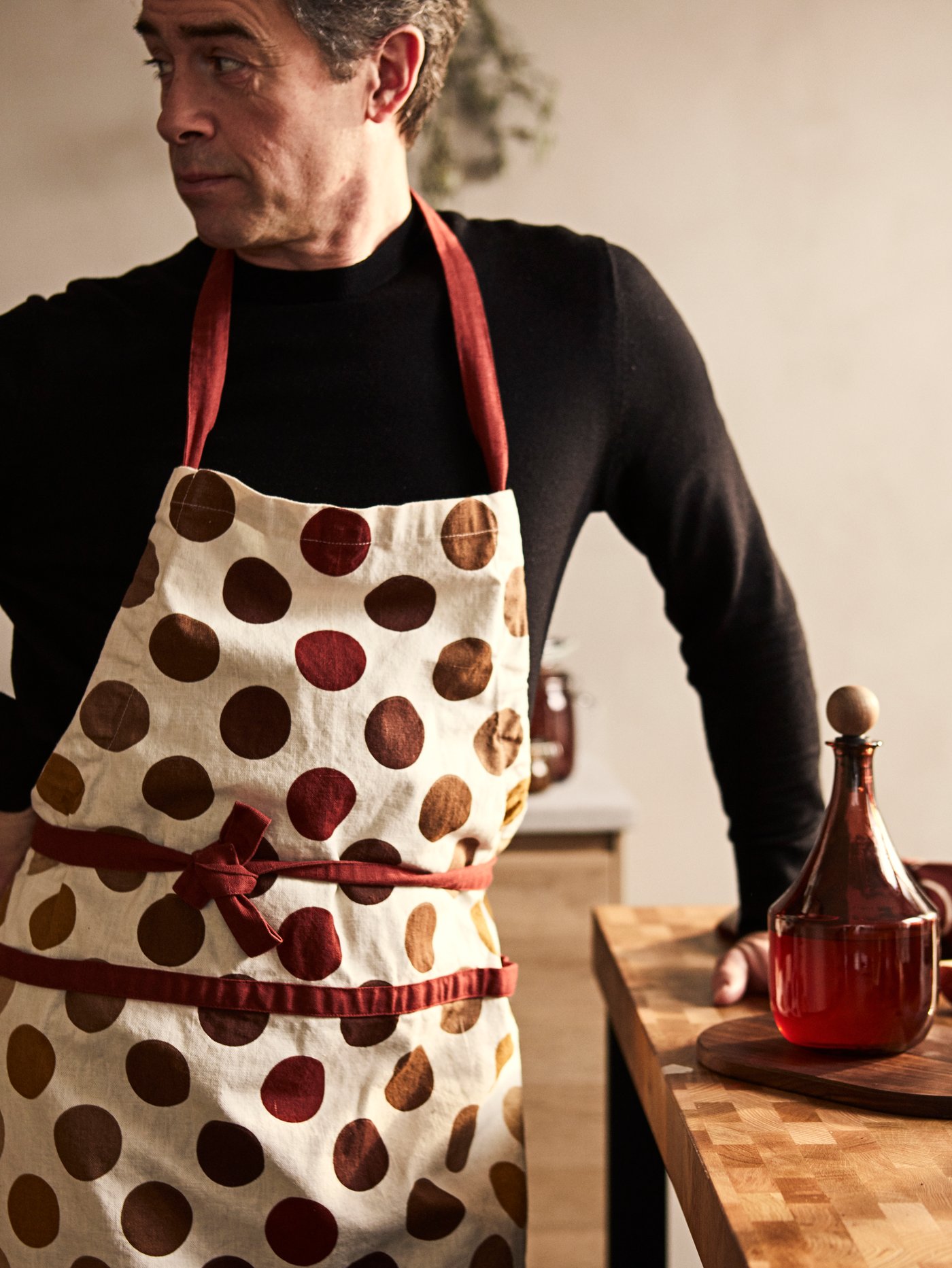 A man is standing in the kitchen wearing a KRÖSAMOS apron. On the countertop next to him is a KRÖSAMOS glass bottle.