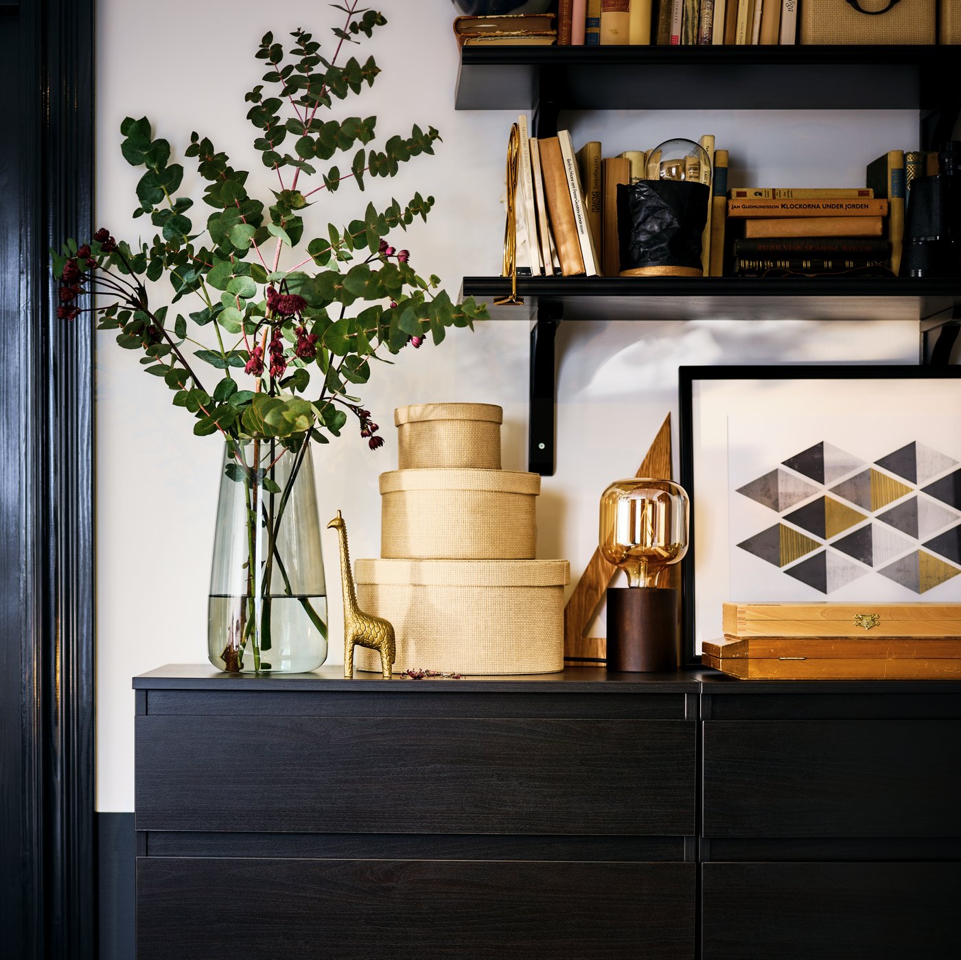 Two KULLEN chests of five drawers with the top filled with decorative objects. Dark, wall-mounted shelves above.