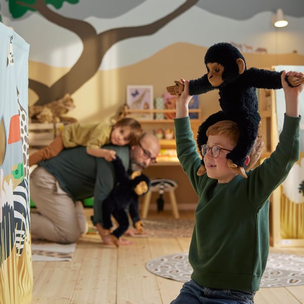 Enfant agenouillé dans une pièce décorée sur le thème de la savane, avec un chimpanzé en peluche SANDLÖPARE sur ses épaules.
