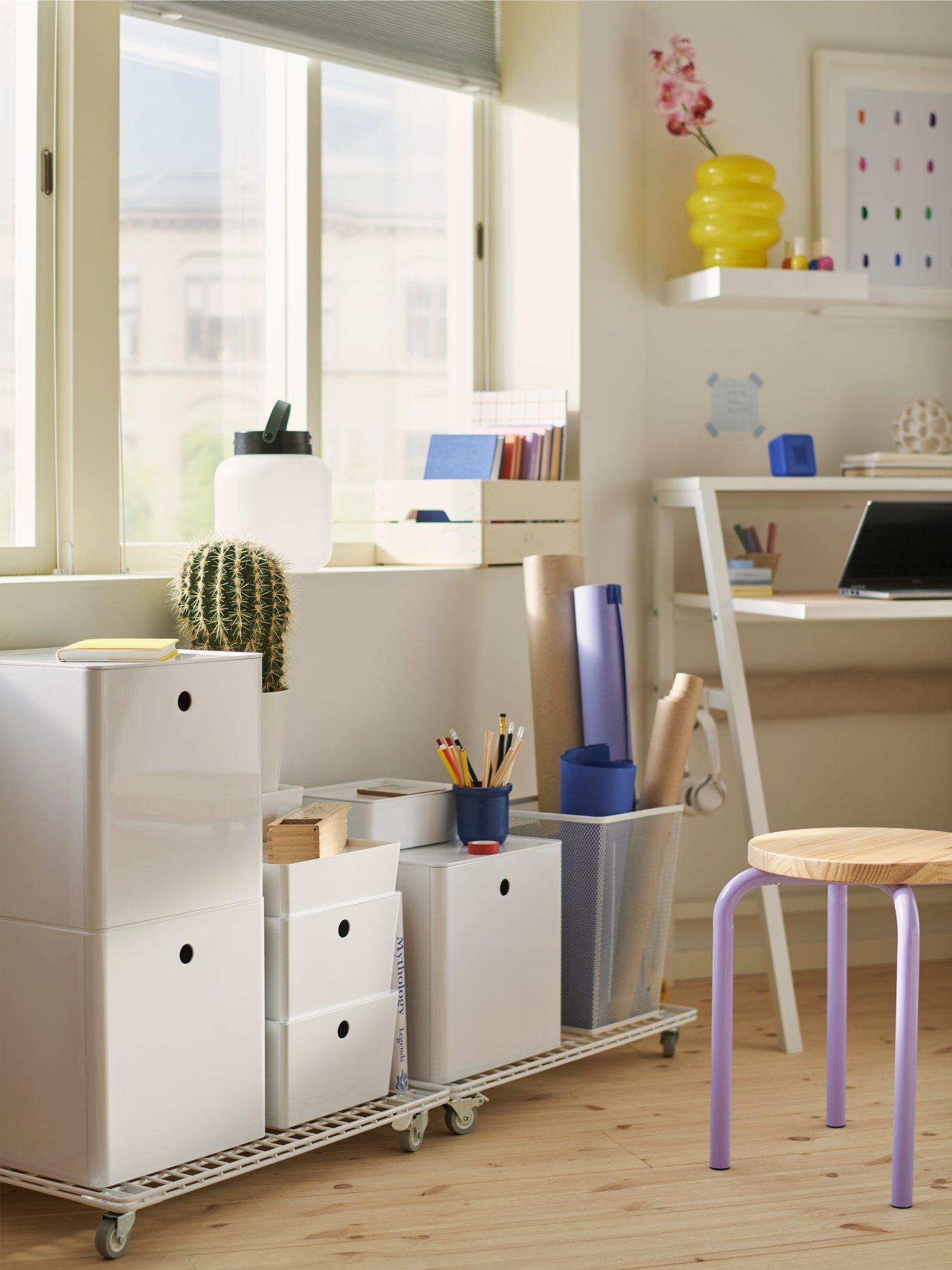 A small home office corner with a window and KUGGIS boxes with lids stacked on two white INVALLNING trolleys for boxes.