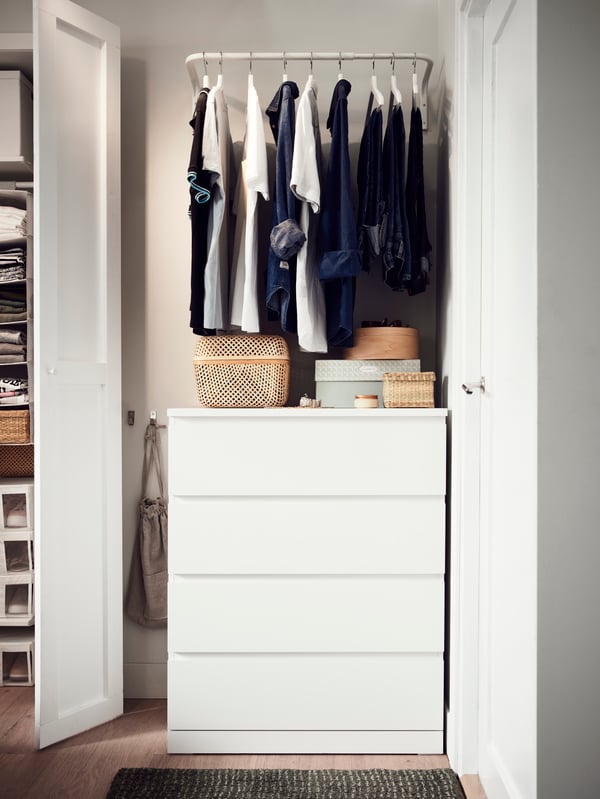 A white MALM chest with 4 drawers with storage boxes on top. A clothes bar above it holds shirts and jeans on hangers.