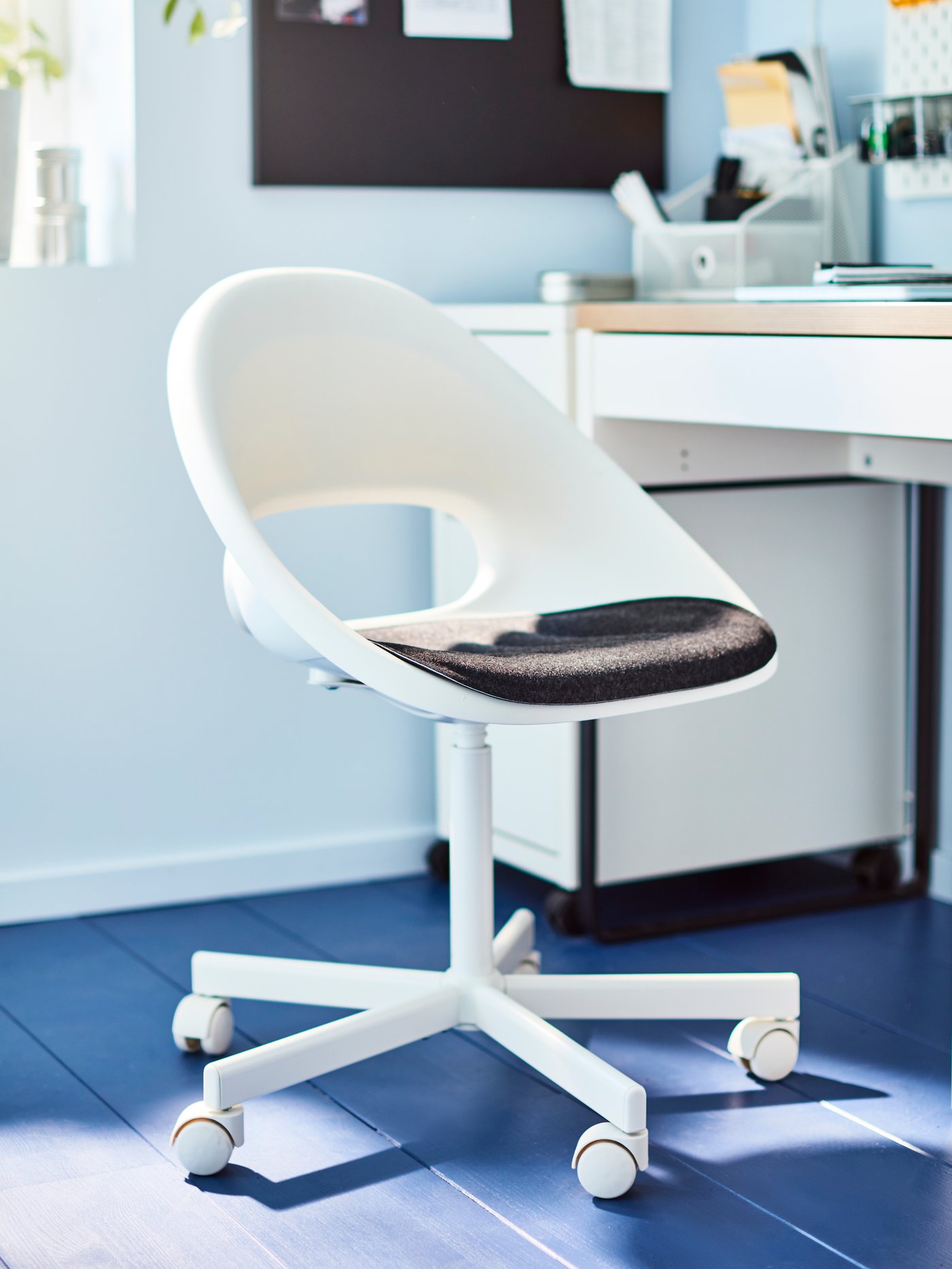 A white swivel chair with dark grey seat pad stands on a blue floor next to a white desk.