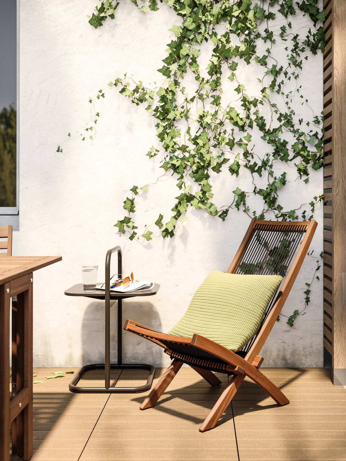 Una silla de madera BROMMÖ para terraza situada al lado de una mesa lateral HUSARÖ y frente a un muro blanco con una enredadera. Se sitúa sobre un piso de deck de madera y tiene sobre ella un cojín verde claro SÖTHOLMEN.
