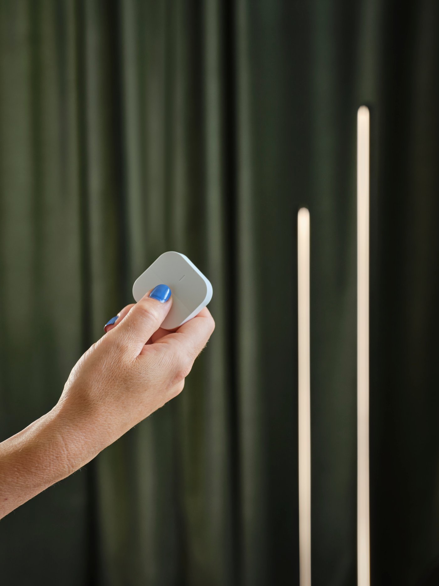 A hand holding a white TRÅDFRI wireless dimmer with a PILSKOTT LED floor lamp and SANELA room darkening curtains behind it.