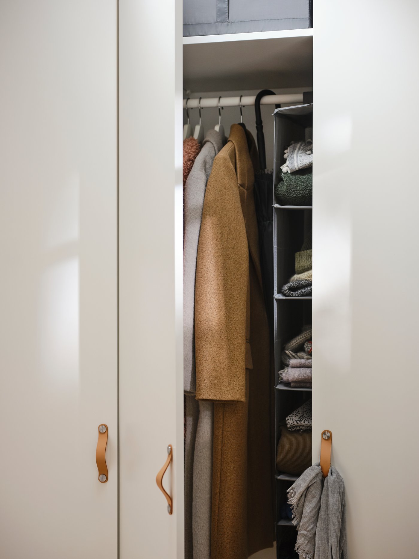 Une armoire ouverte avec poignées en cuir dévoile des vêtements suspendus, dont un manteau brun, et des étagères à droite avec des vêtements pliés et divers objets.