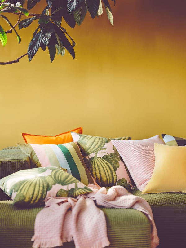 PARKSALLAT cushion covers with clashing patterns sit on top of a JÄTTEBO sofa, in front of a mustard coloured wall.
