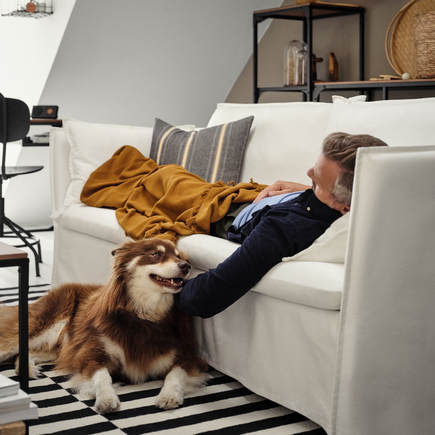 Une personne est allongée sur un canapé blanc dans un salon et caresse un chien couché sur un tapis rayé.