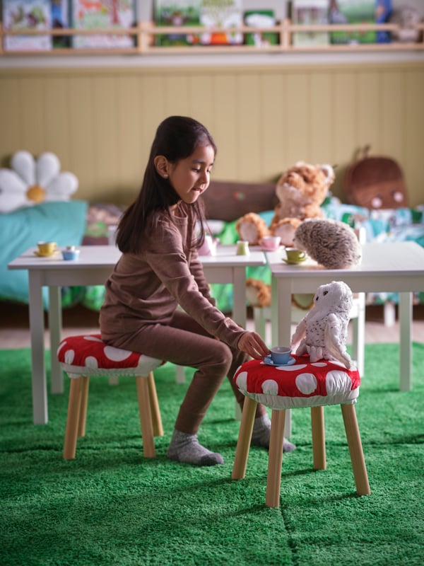 A child is sitting on a stool with a SKOGSDUVA toadstool-shaped stool cover playing with a SKOGSDUVA snowy owl soft toy.