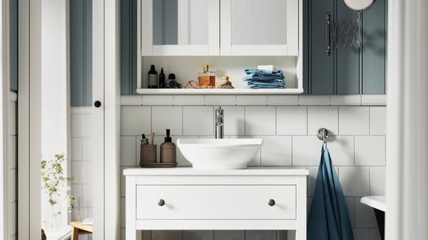 Ett vitt HEMNES öppet tvättställ med en låda är placerat i ett vitt och blått badrum, bredvid ett vitt skåp med spegel.