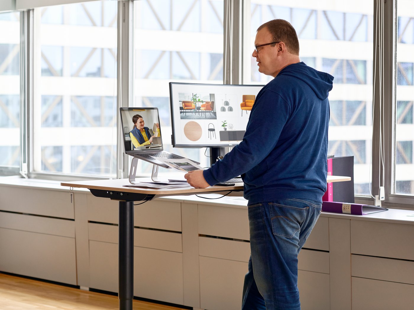 A person is standing in an office at a desk holding two monitors and is talking to a woman on the screen.