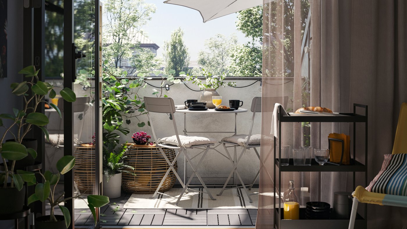 A balcony with a grey SUNDSÖ outdoor table and two grey outdoor chairs on a rug, plus a coffee table, plants and a parasol.