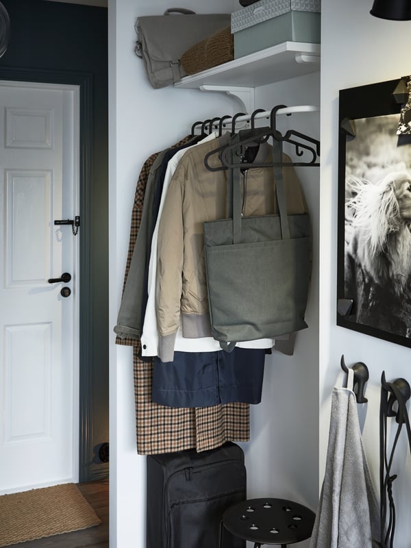 An odd nook with a white clothes bar with jackets and coats hanging, a black cabin bag, a shelf with boxes and bags.