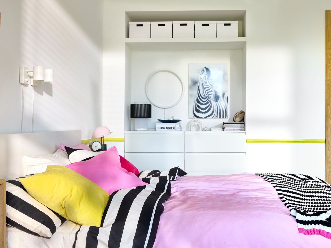 A bedroom with white walls, built-in storage in the back and a double bed with bright pink and black/white striped bedding.