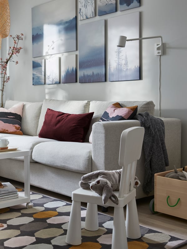 En un living de tonos blancos, grises y celestes, se ve en primer plano una silla infantil MAMMUT blanca y un conejo de peluche posado sobre ella. El living está amoblado con un sillón VIMLE de tres cuerpos y cuadros GRÖNBY de tonos celestes. 
