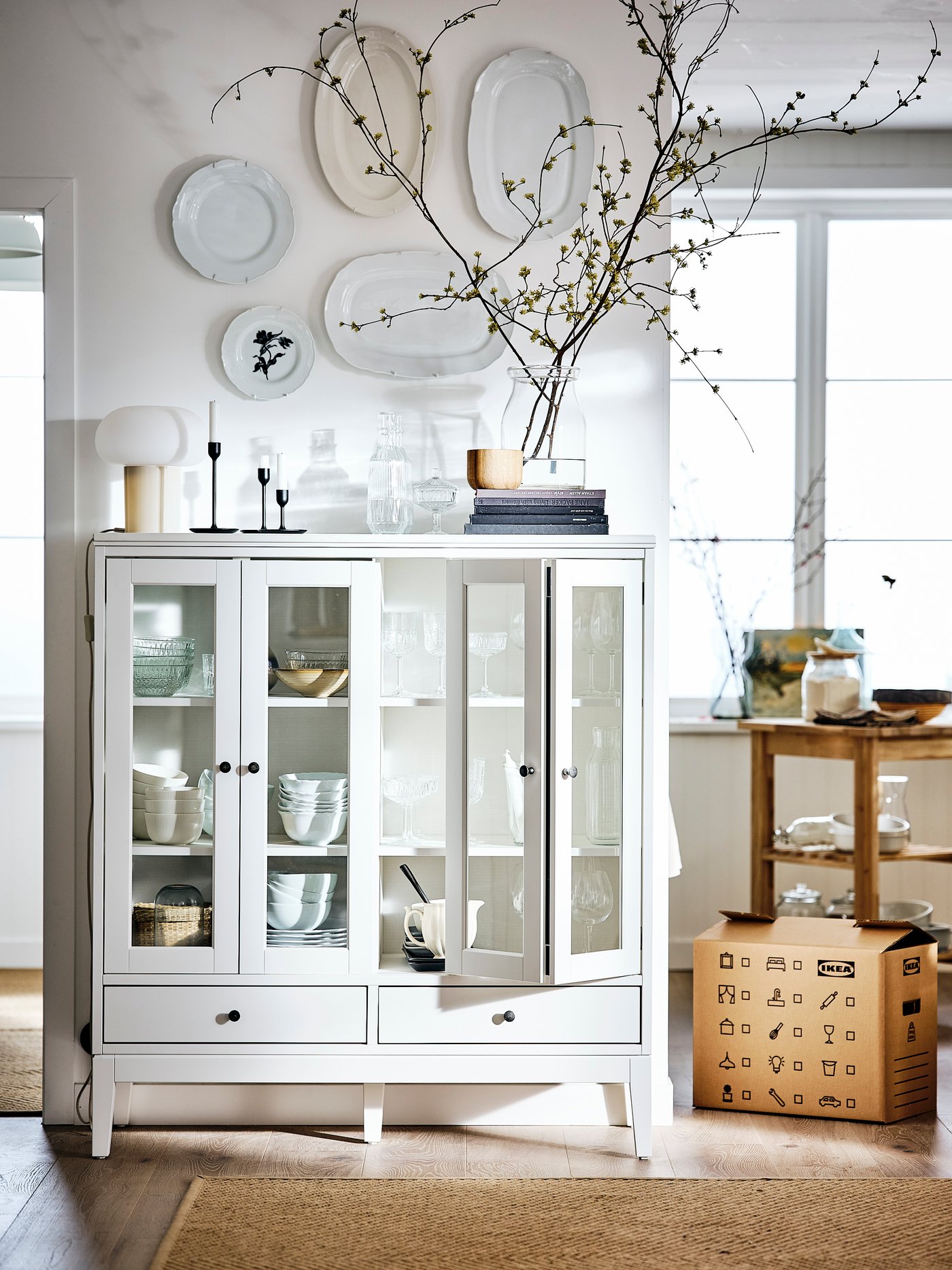 A room with white walls and floors with an IDANÄS display cabinet above which plates are placed on the wall.