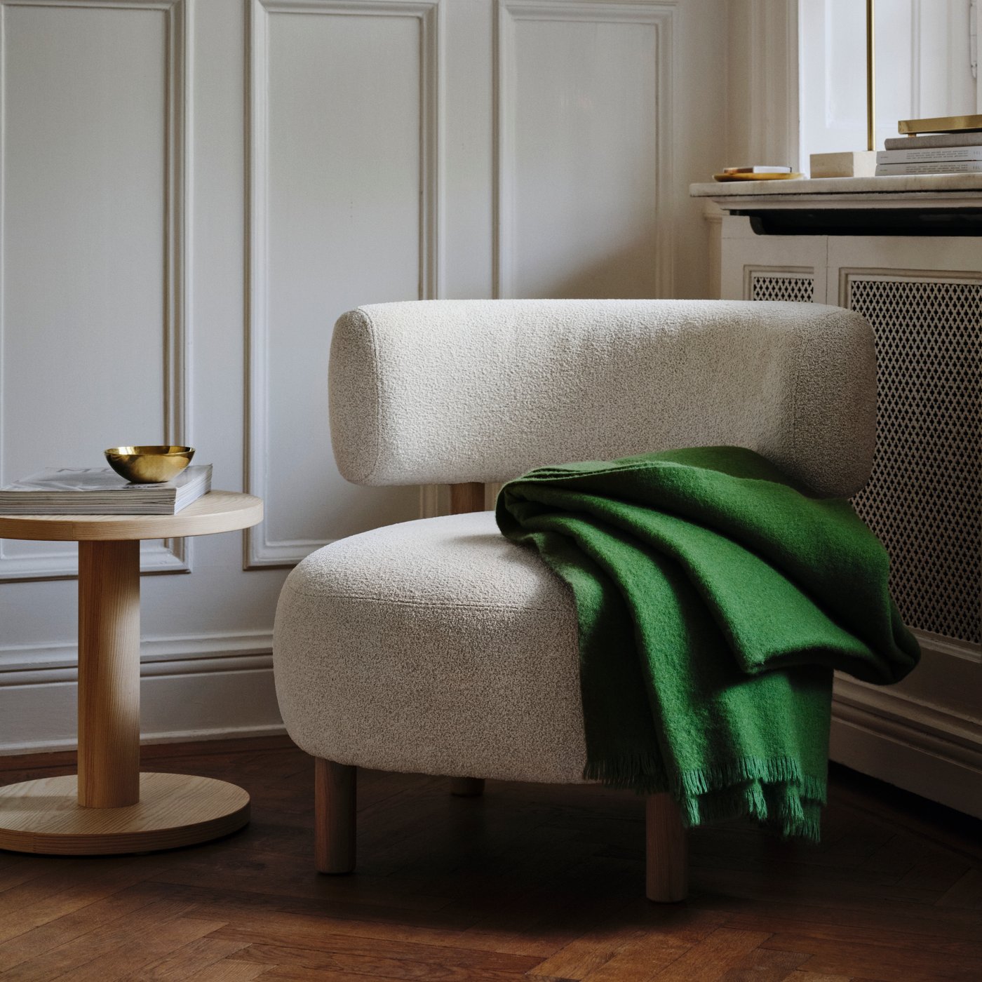 A light living-room corner arranged with a grey-and-white STOCKHOLM 2025 armchair, green throw, and pine-veneer side table.