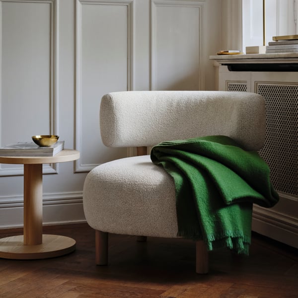 A light living-room corner arranged with a grey-and-white STOCKHOLM 2025 armchair, green throw, and pine-veneer side table.