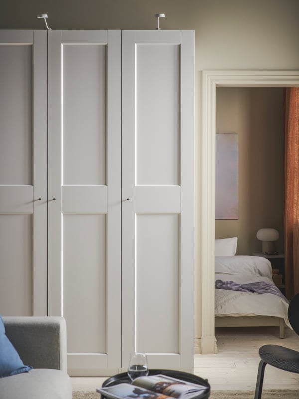PAX/GRIMO wardrobe outside a bedroom. White doors and NYDALA knobs. Outside the bedroom there is a grey VIMLE sofa bed.