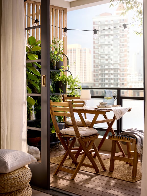 A small balcony has a light brown stained acacia ASKHOLMEN foldable table and 2 folding chairs by potted plants on a side wall.