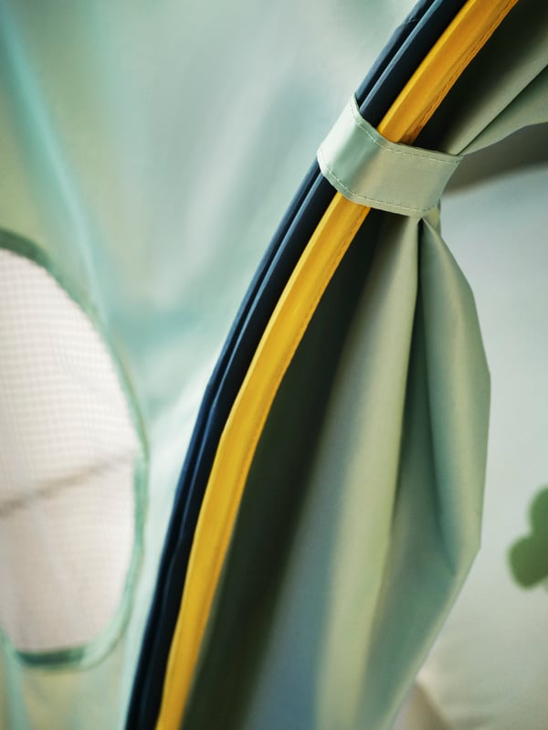A close-up of the edge of the blue/green ELDFLUGA bed tent with yellow piping and a small green strap holding the tent open.