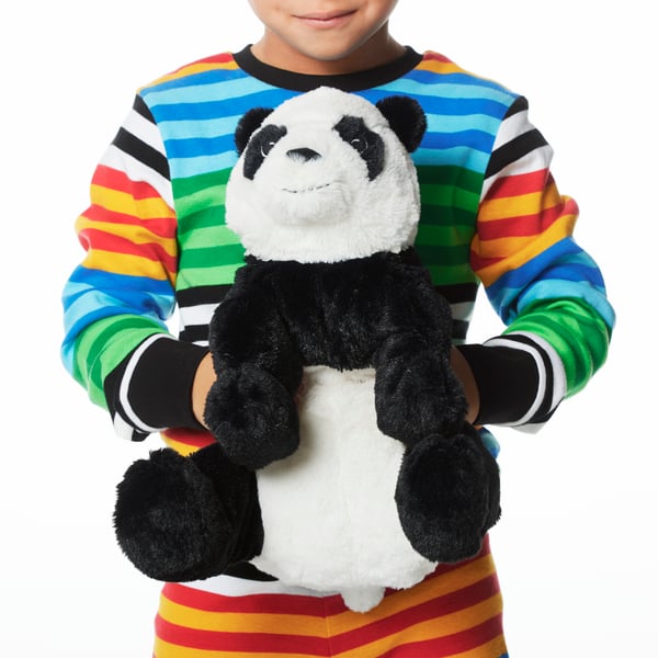 A child in colourful striped pyjamas holds a KRAMIG soft toy panda bear in front of their torso.