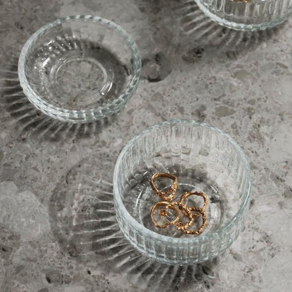 Detalle de un recipiente de vidrio SILVTJÄRN con tapa sobre una mesa gris con pequeñas joyas dentro.