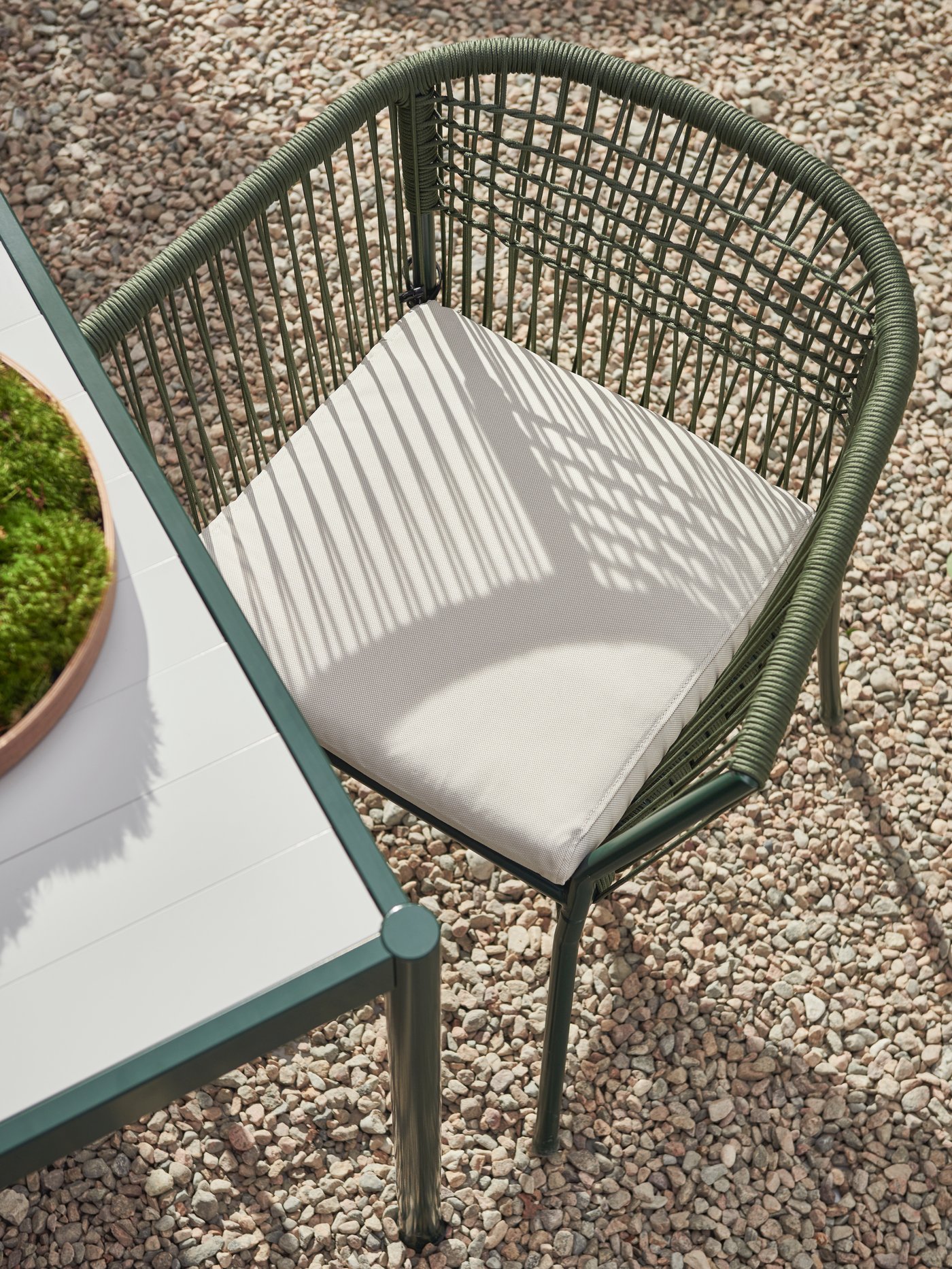 Grüner Gartenstuhl mit Sitzpolster neben einem weißen Tisch auf Kiesboden.