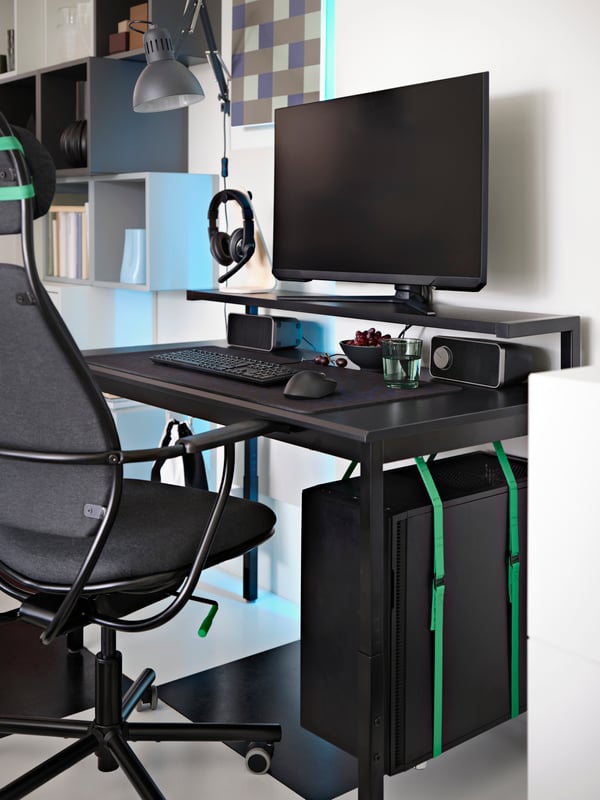A black UTVSNING gaming desk with shelf has a monitor, keyboard, mouse, etc. on it and a CPU held below using green straps.