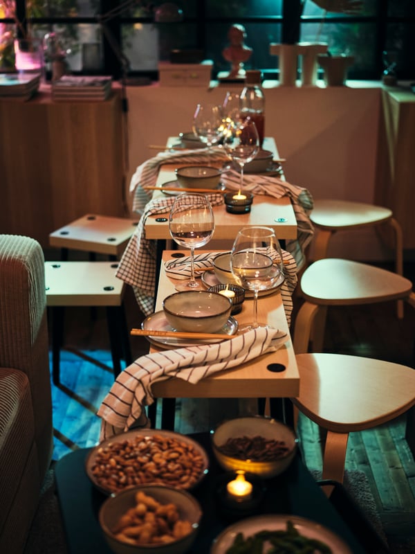 A living room set up for guests and lights dimmed, IKEA FRIDNÄS tables with birch tools set up for dinner.