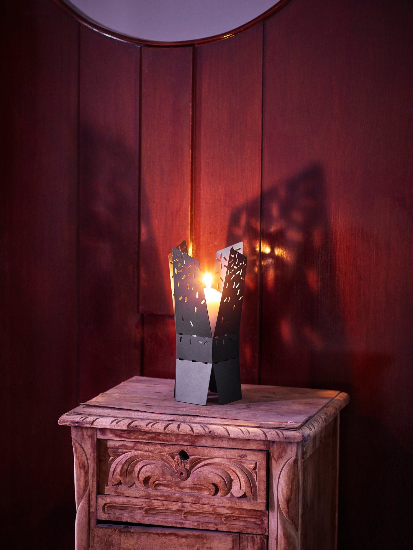 A large candle burning inside a black ÖMSESIDIG candle holder in powder coated steel on a wooden side table with carvings.