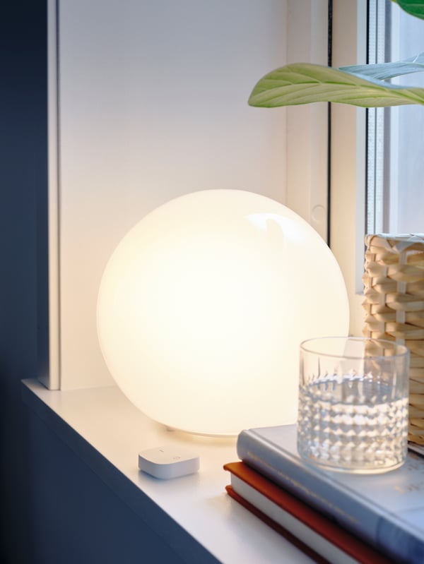 A lit FADO table lamp on a windowsill near a TRÅDFRI wireless dimmer and a FRASERA whiskey glass on top of some books.