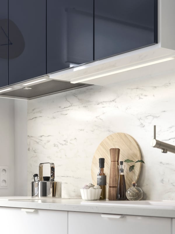 IKEA UNDERVERK kitchen hood is integrated under black-blue cabinets with lit halogen bulbs above a marble effect worktop.