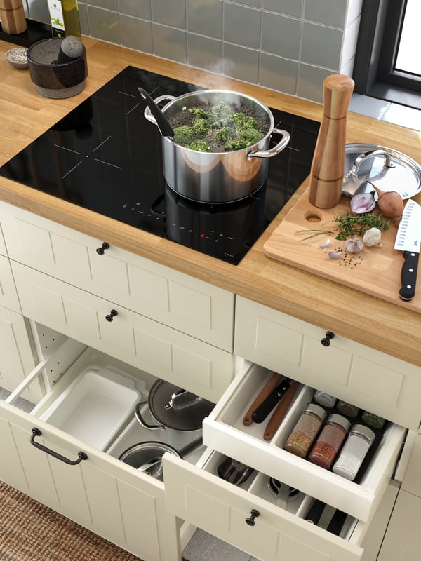 Une casserole IKEA 365+ avec couvercle sur une plaque de cuisson à induction noire, avec en dessous des tiroirs ouverts contenant des casseroles, des plats et des épices.