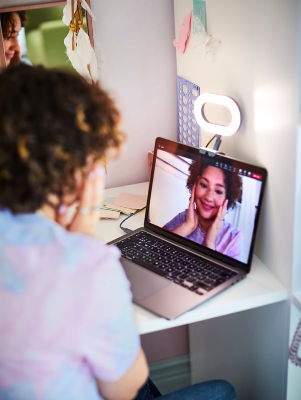 A young person facing a laptop with a STÄNKREGN dimmable ring light on it. A pink GRANVÅG mirror is on the wall beside them.