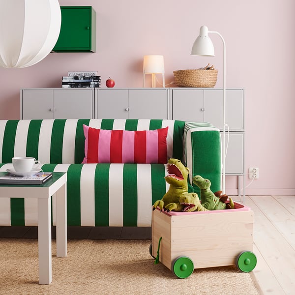 A living room setting with a green and white striped couch decorated with a pink striped pillow. In front of the couch sits a wooden storage box with green wheels. 