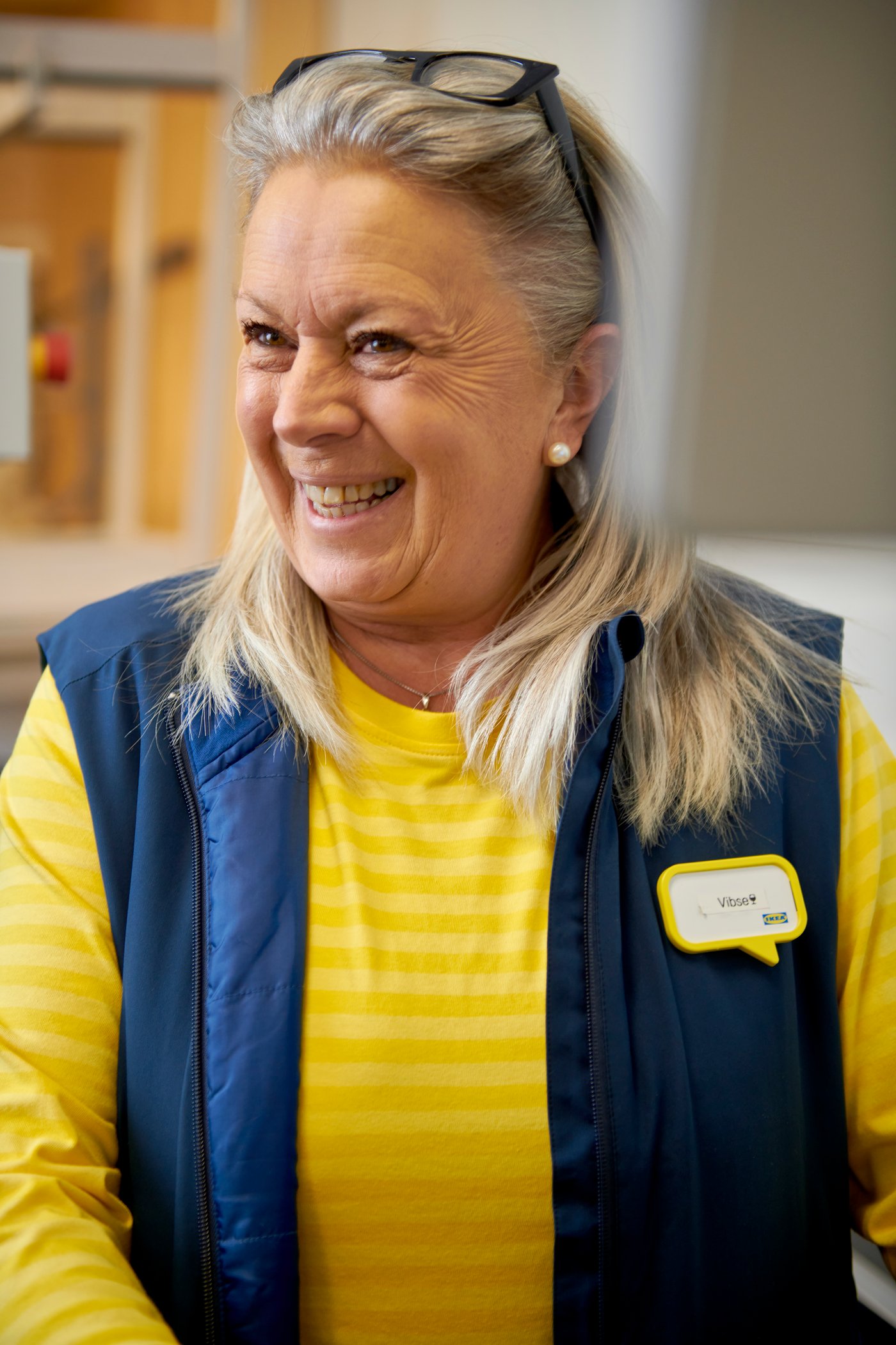 IKEA medewerker in een geel IKEA shirt en blauw vest lacht, terwijl haar bril bovenop haar hoofd staat.