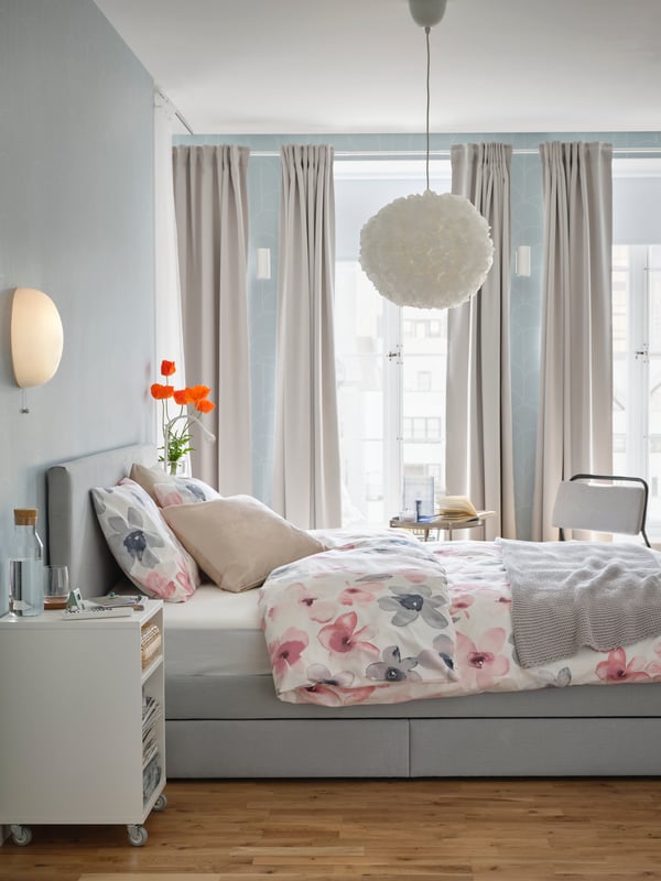 Il lato di un letto GLADSTAD con biancheria da letto floreale e una lampada a sospensione sopra, di fronte a una finestra con tenda - IKEA